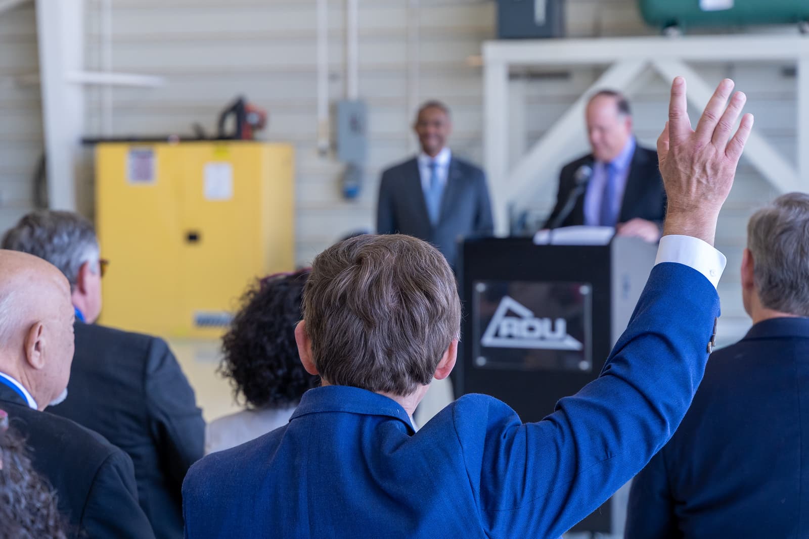 Person in blue suit raising hand during business presentation with speakers at podium and yellow cabinet in background.
