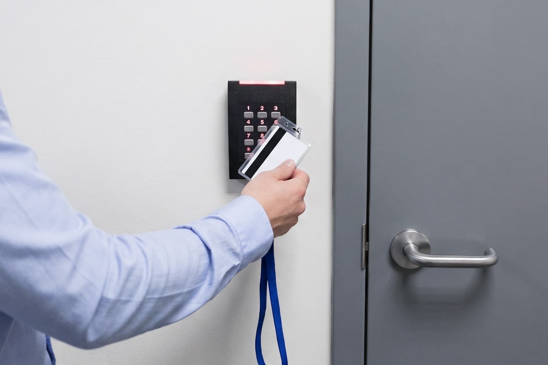 Person in light blue shirt holding white access card to black keypad reader mounted on white wall next to gray door.