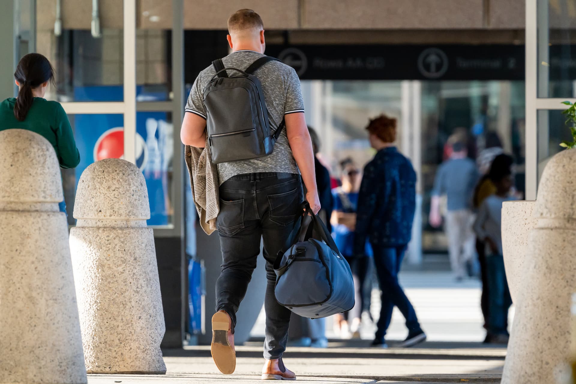 Man with backpack and duffel bag walking toward airport entrance with people in background.