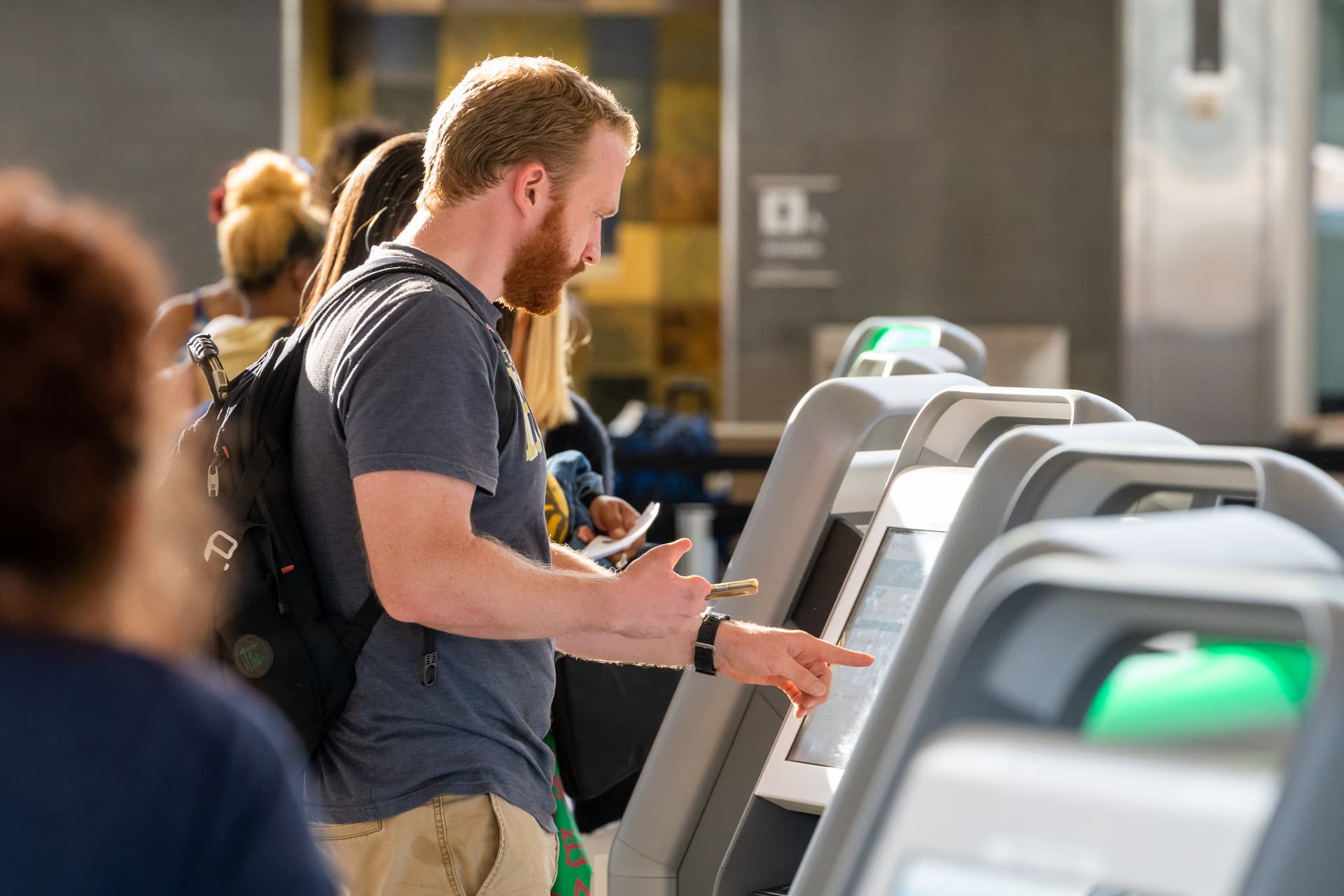 Man with backpack using self-service kiosk with green screen display in modern indoor facility.