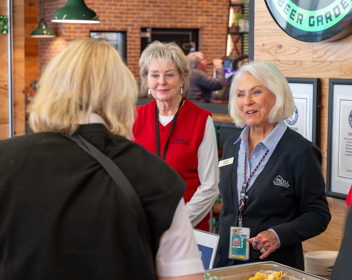 Two women in branded vests greet a visitor at a welcome desk in a modern space with brick walls and green pendant lights.