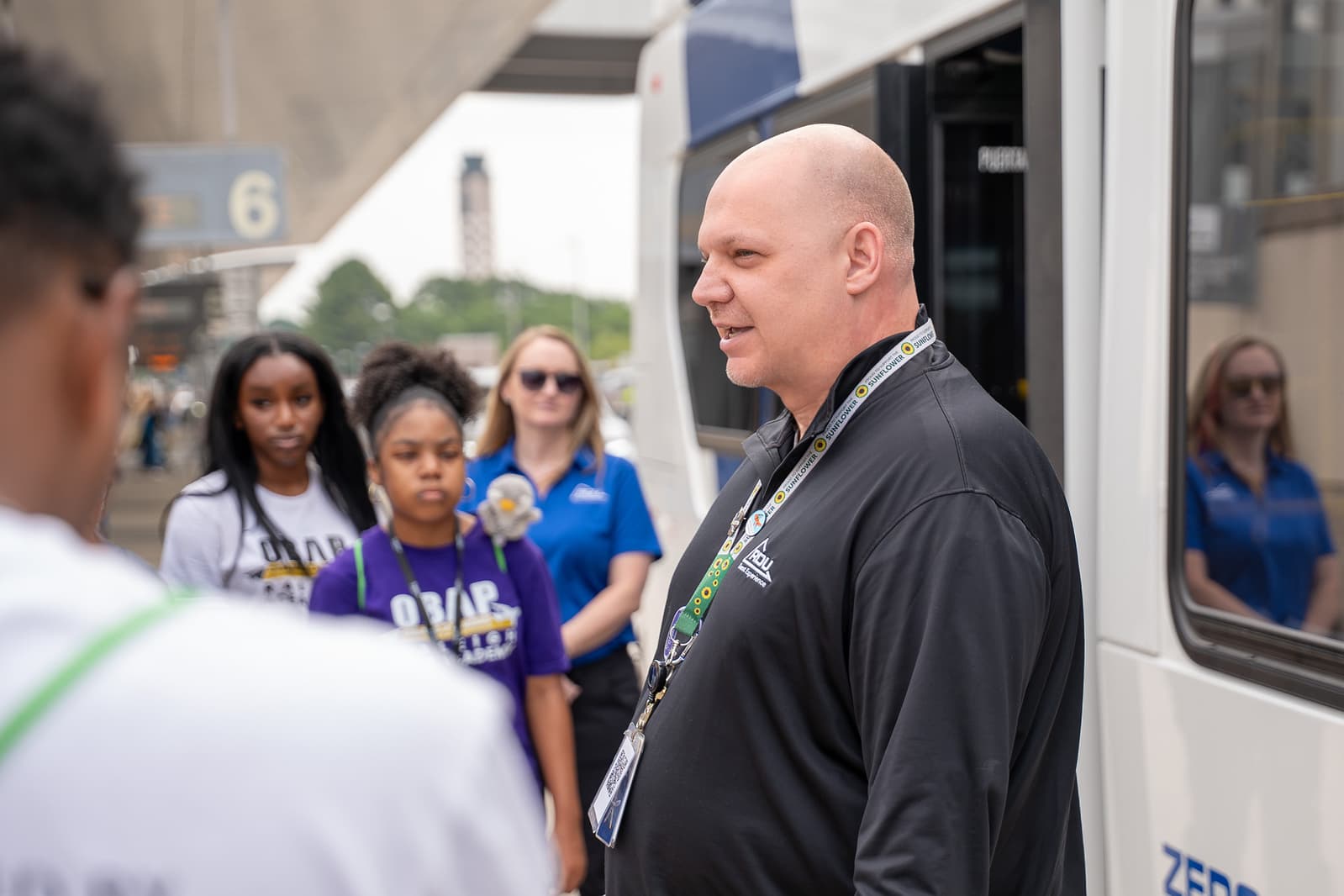 An RDU Guest Experience employee in black shirt speaks to a group of students near an airport shuttle bus.