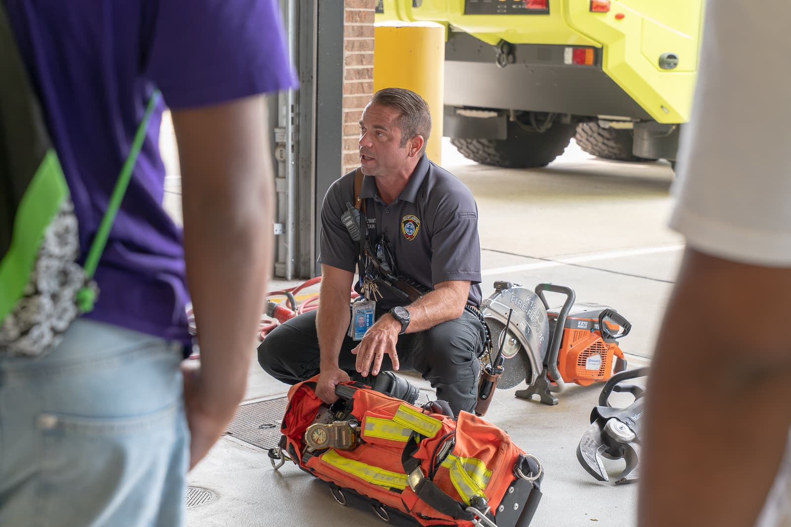 An RDU firefighter in gray polo shirt, kneeling beside emergency equipment, demonstrates tools to visitors at the airport fire station.