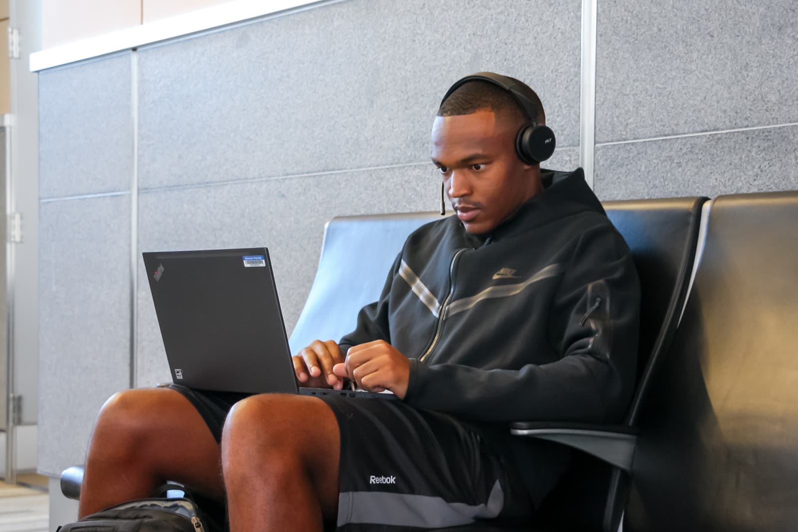 A guest wearing headphones and a hoodie works on a laptop while sitting near the airport gate.