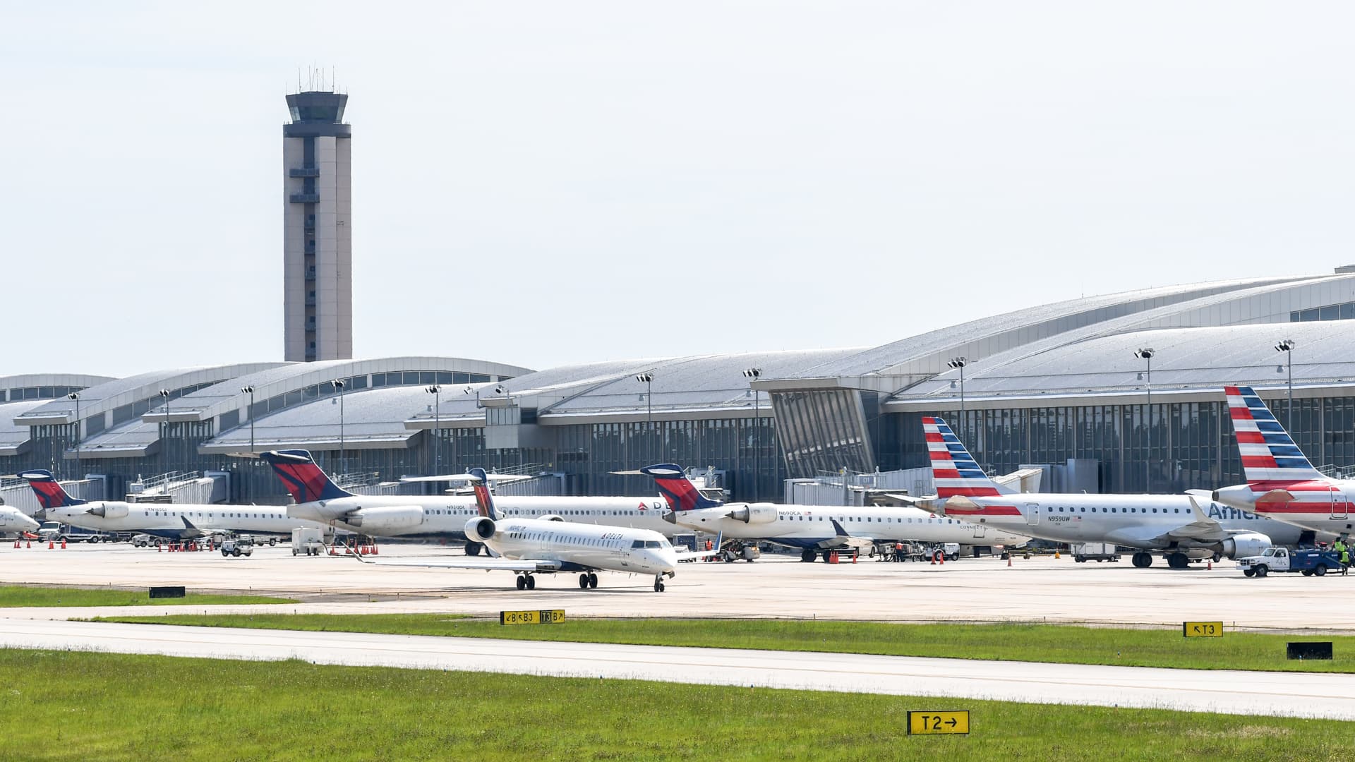 Multiple commercial aircraft parked at airport gates with a modern curved terminal building and control tower in background.