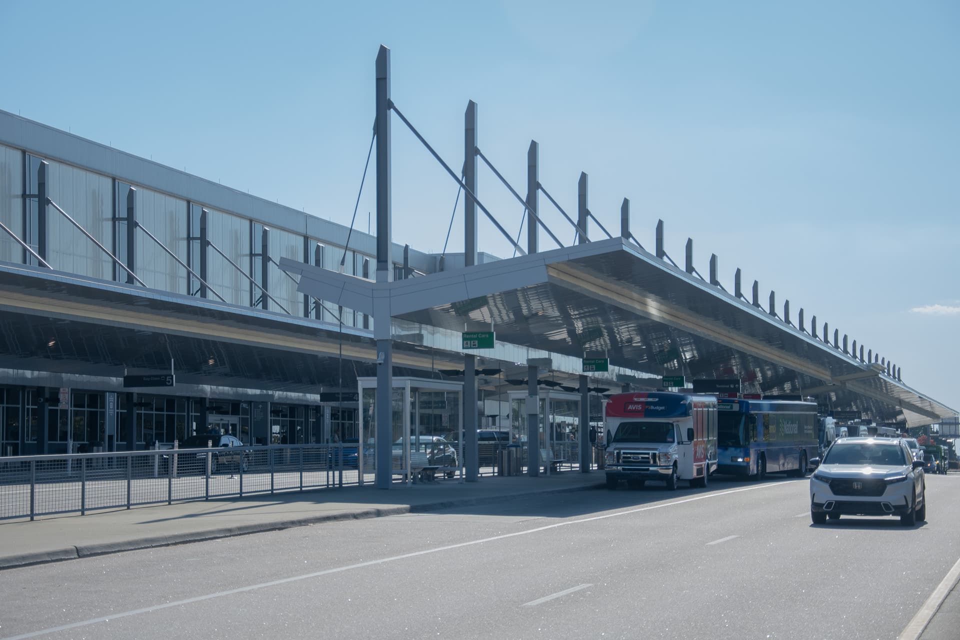 Modern airport terminal with distinctive angled support columns and cable-stayed canopy over roadway with buses and vehicles.