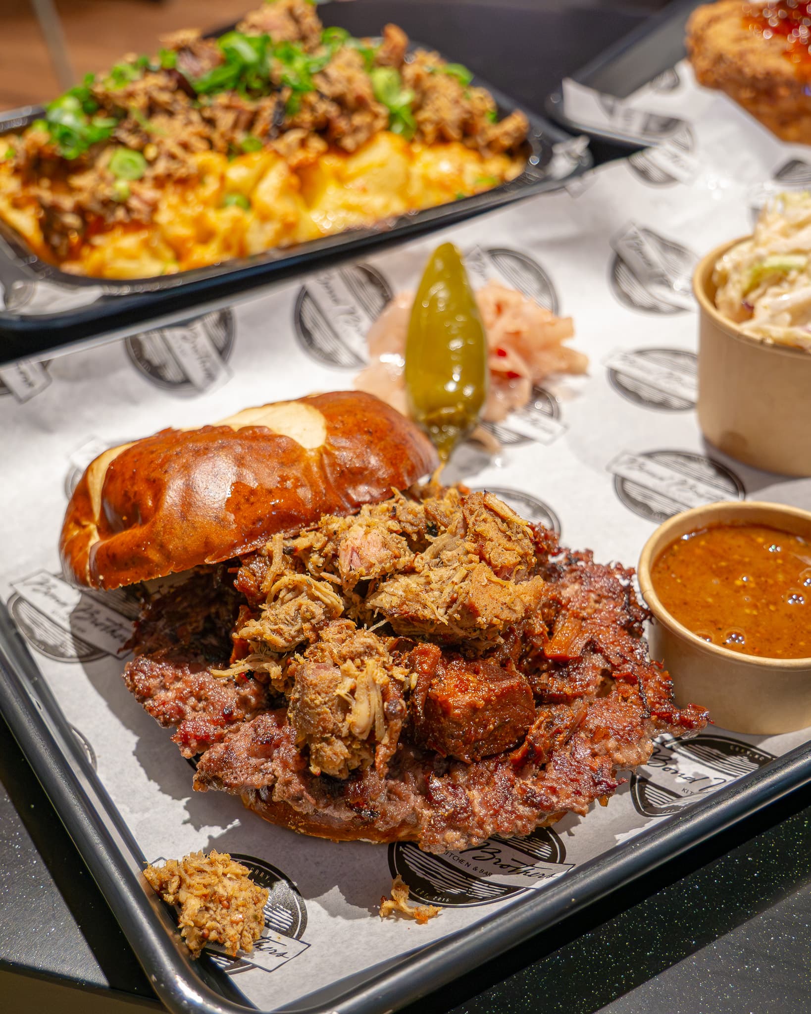 Crispy fried chicken sandwich on pretzel bun with pulled pork, pickles, and sauce on tray with mac and cheese in background