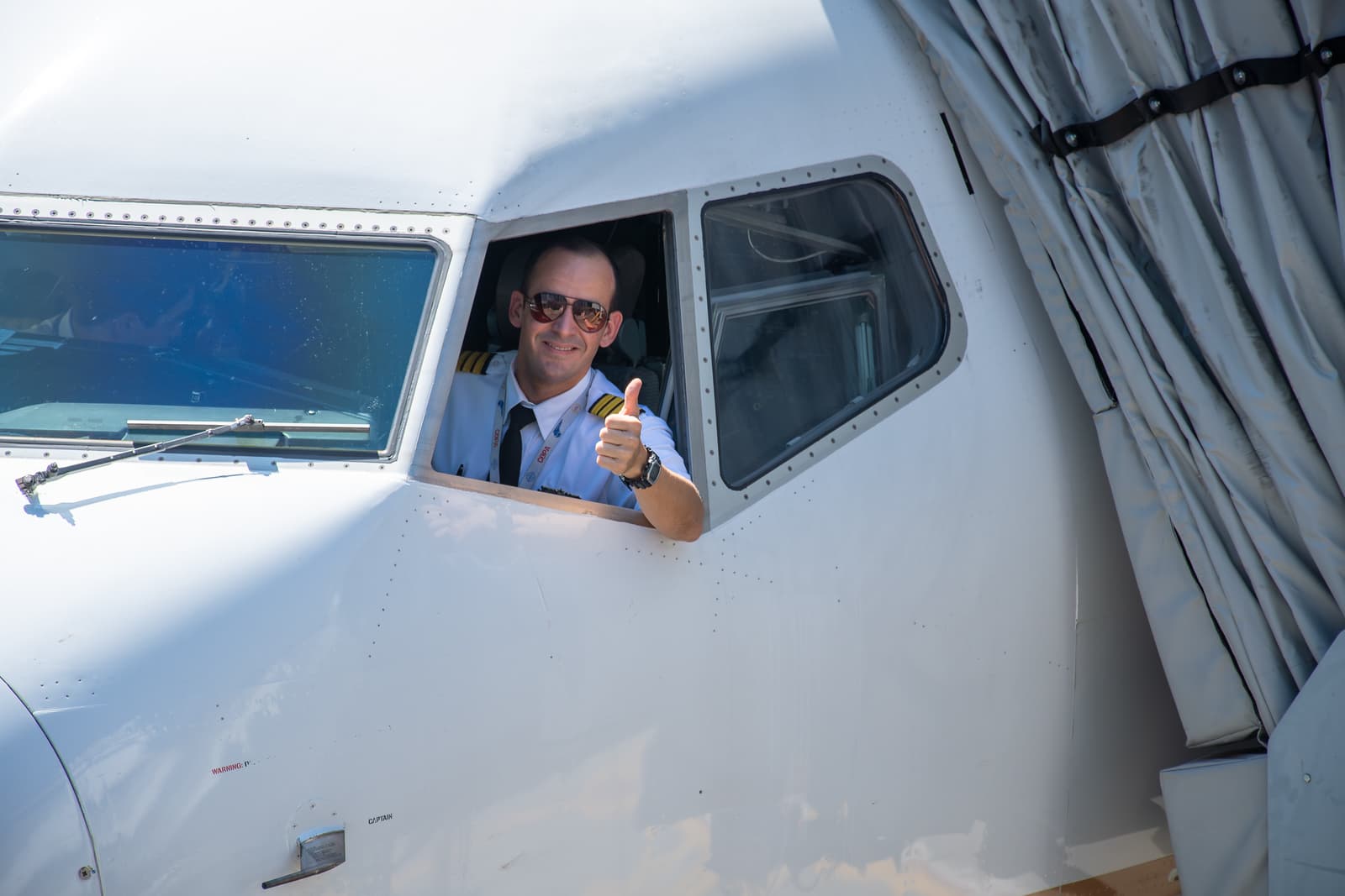 Smiling pilot in sunglasses giving thumbs up from aircraft cockpit window wearing white uniform with gold stripes.