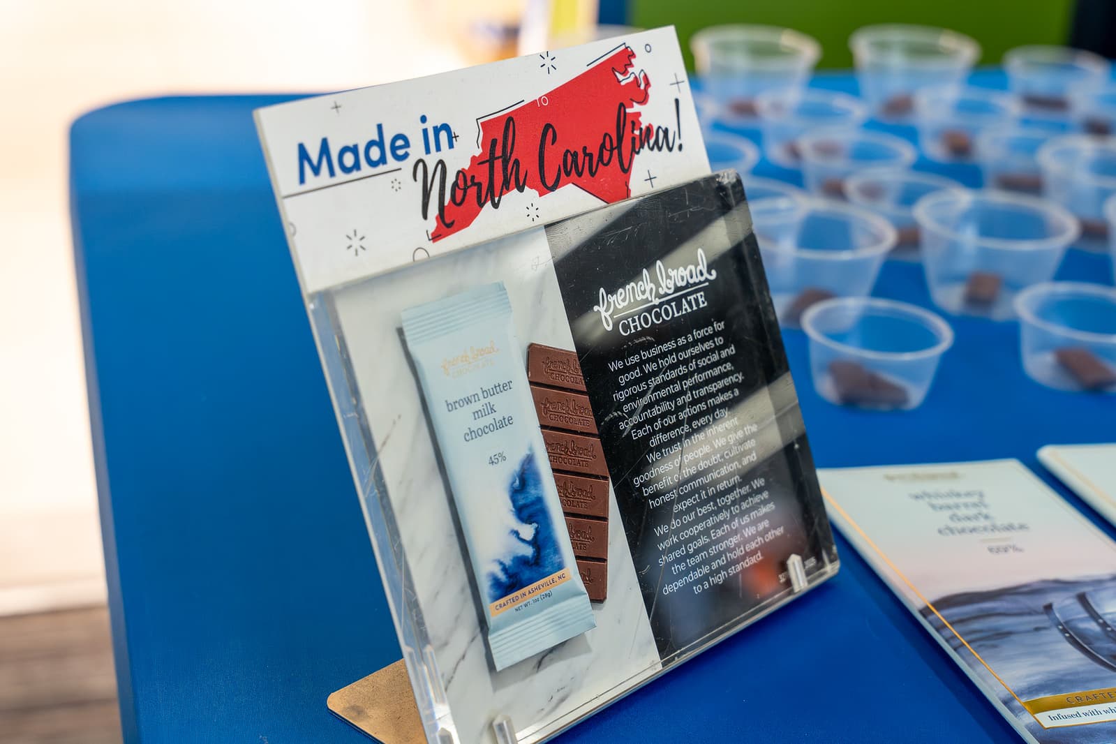 Made in North Carolina sign with red state outline displayed on blue table with chocolate samples and clear cups