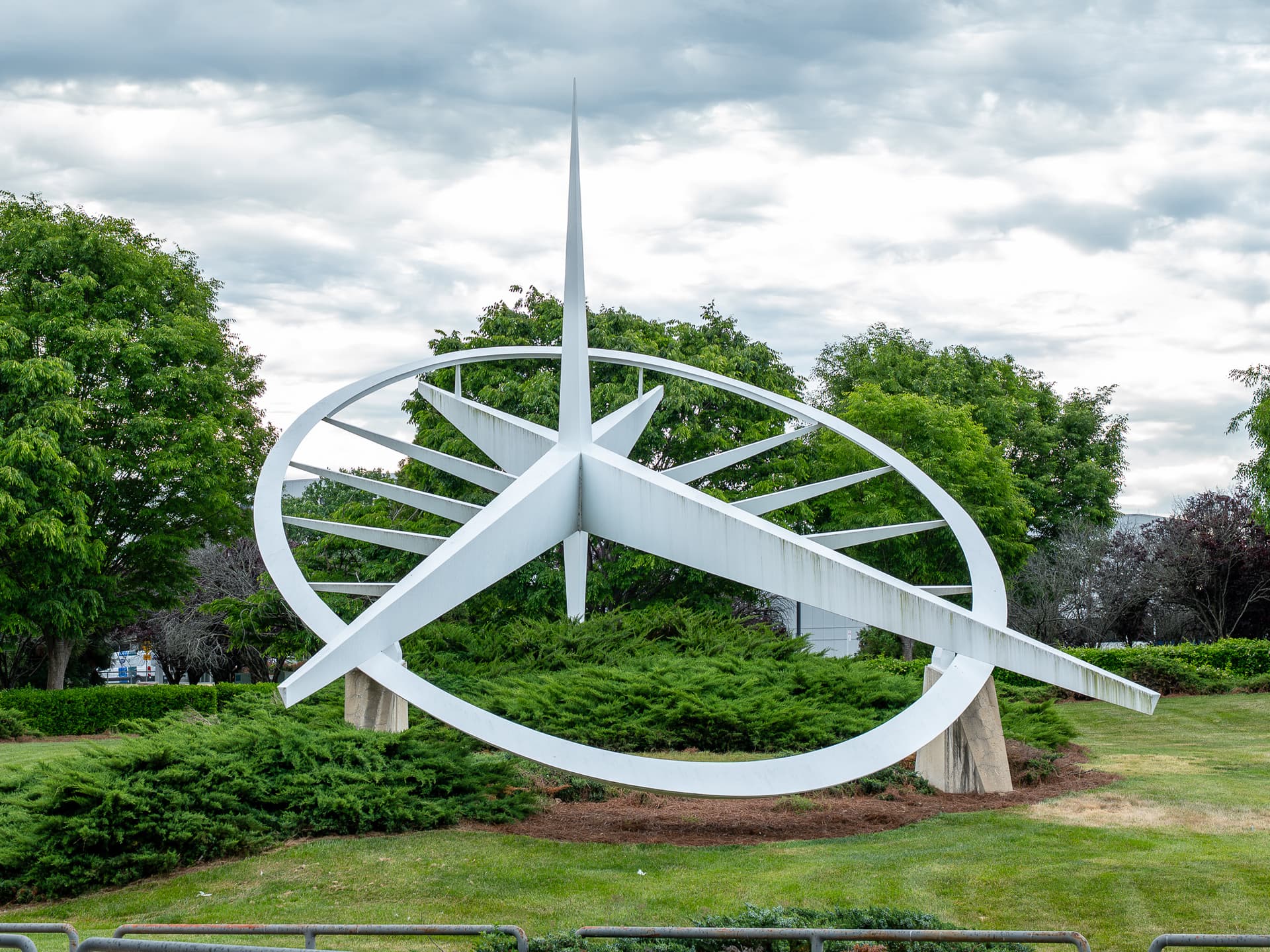 Modern white metal sculpture with circular and angular elements on green lawn with trees and cloudy sky background.