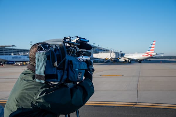Cameraman filming on airport tarmac with American Airlines plane in background under clear blue sky.