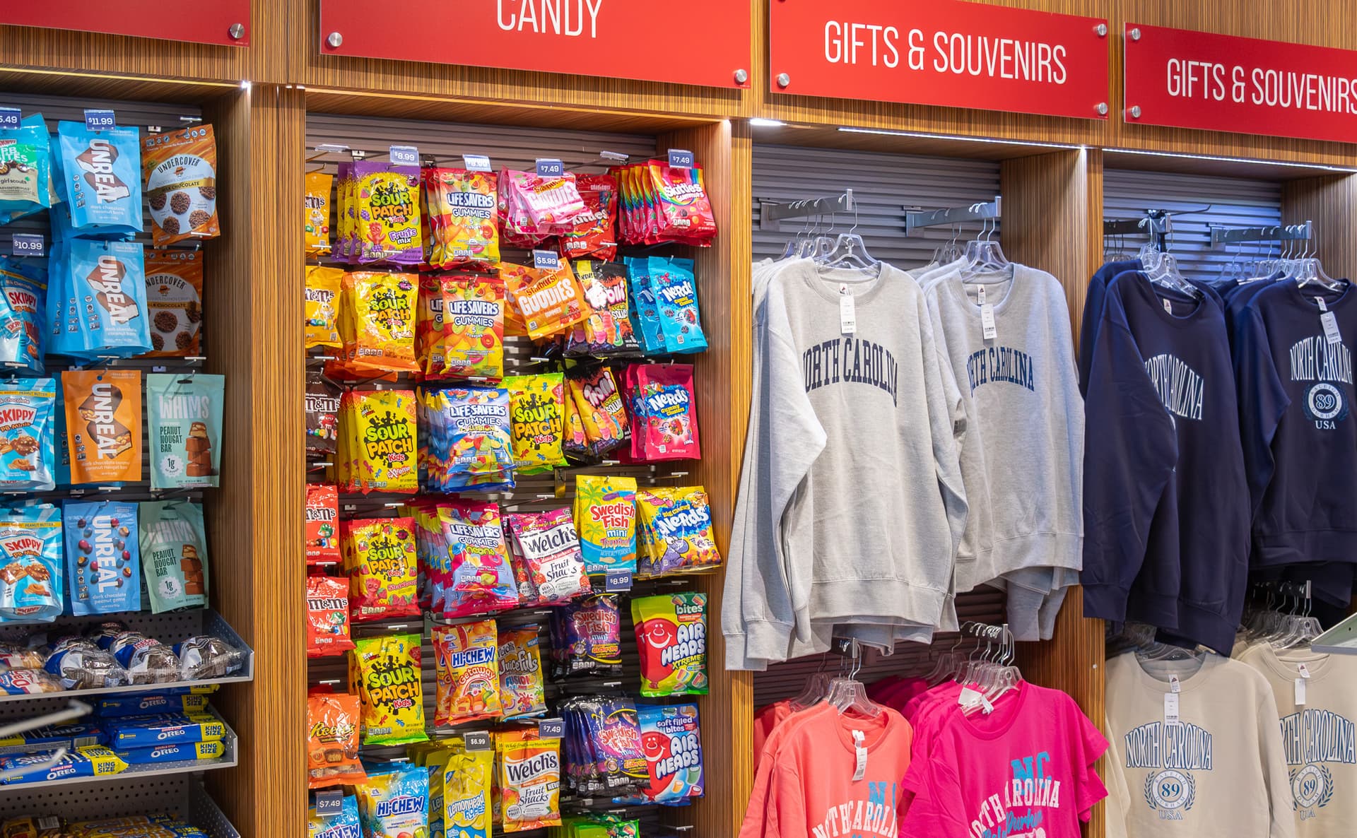 Store display with colorful candy packages on left and North Carolina branded sweatshirts on right under red signage.