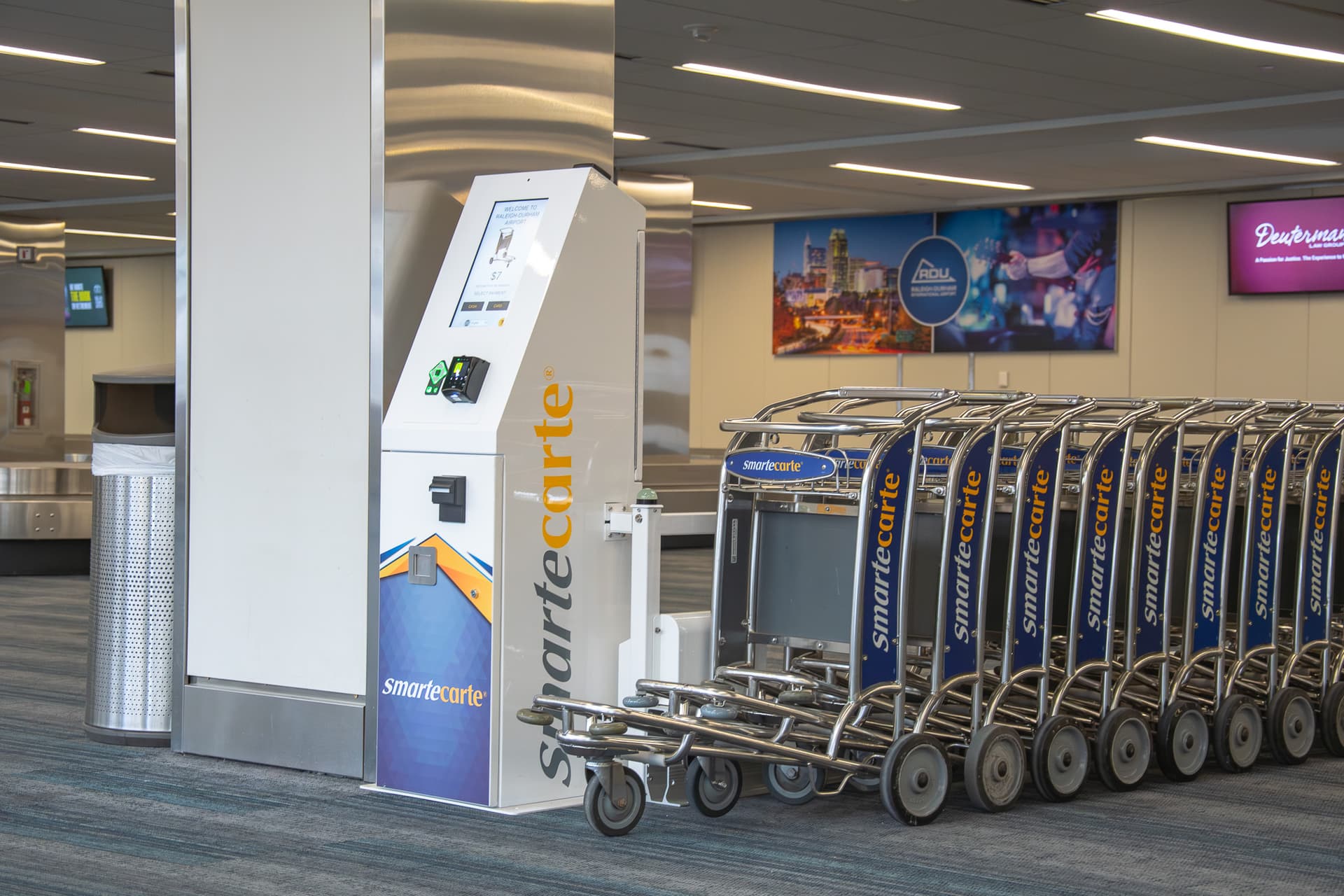 Airport terminal with smartecarte kiosk and row of luggage carts near column with overhead lighting and wall displays.