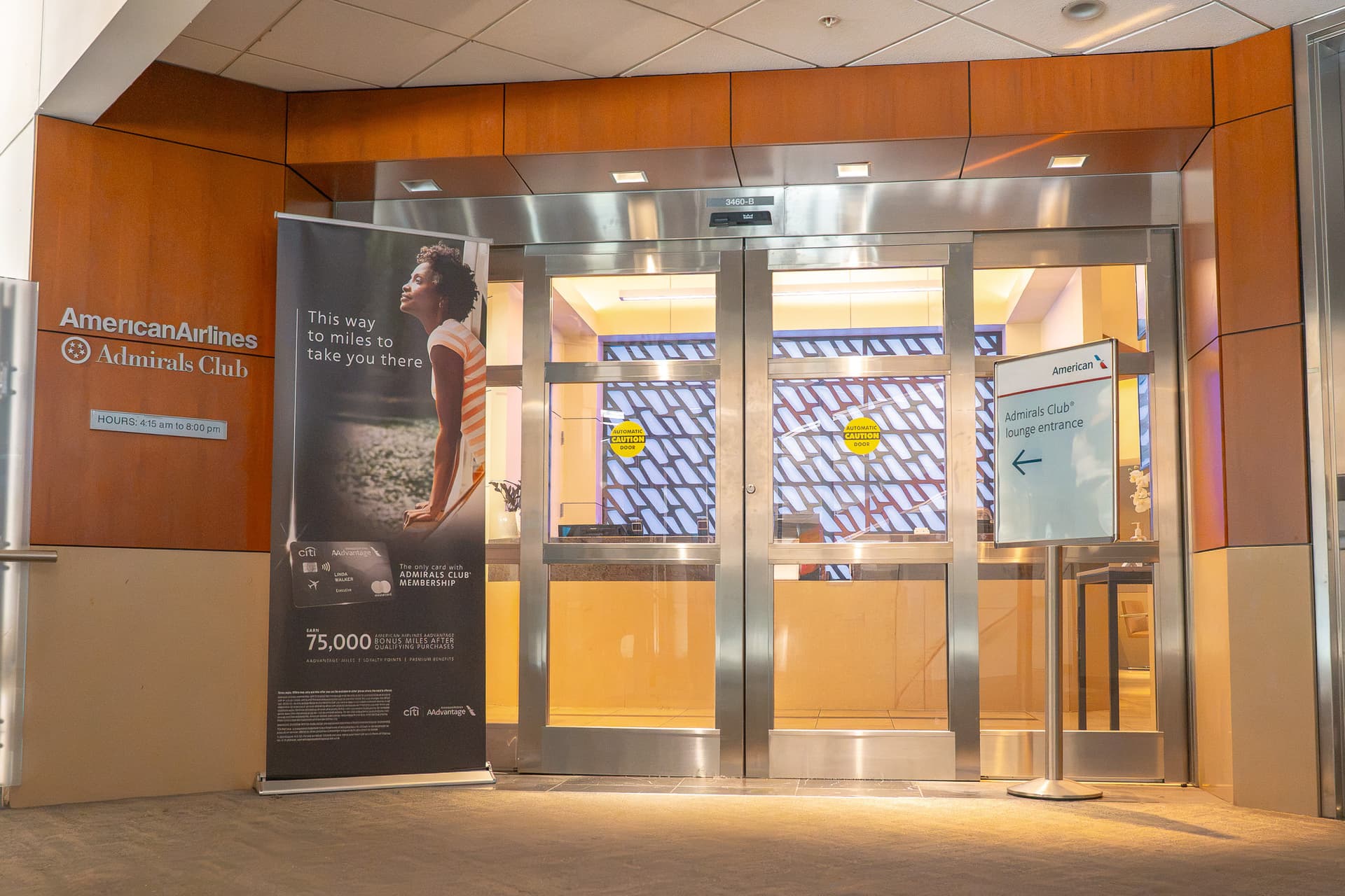 American Airlines Admirals Club entrance with wood paneling, glass doors and promotional banner in airport terminal.