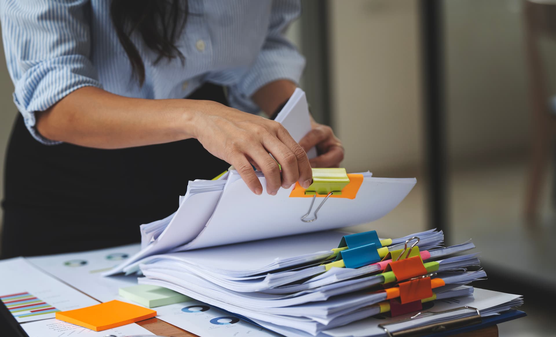 Person in blue striped shirt organizing stacks of white papers with colorful binder clips and sticky notes on desk.