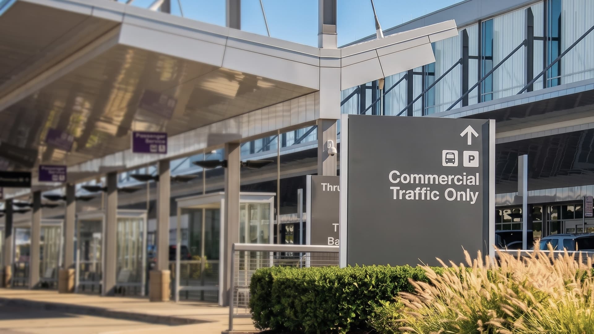 Modern airport terminal with glass facade and Commercial Traffic Only sign near parking structure under blue sky.