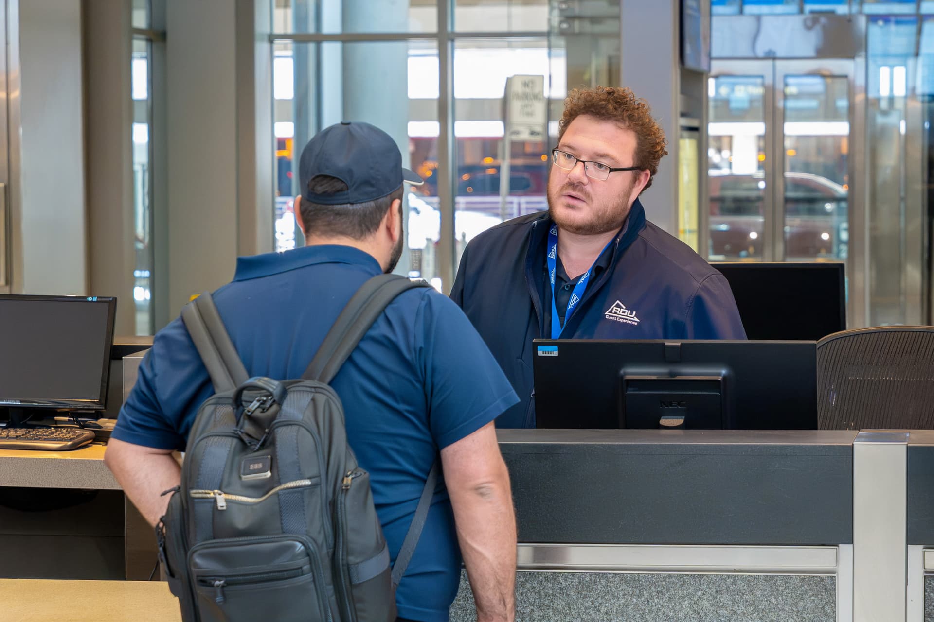 Staff member in navy uniform with glasses assists customer with backpack at service counter in modern facility.