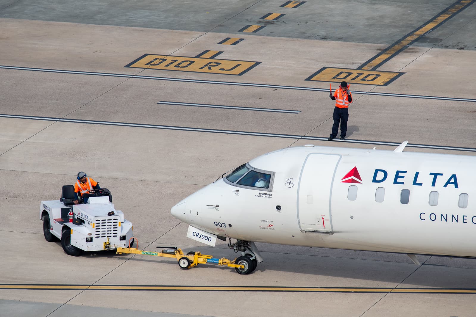 Ground crew in orange vests guiding a Delta Connection aircraft on tarmac with yellow runway markings visible.