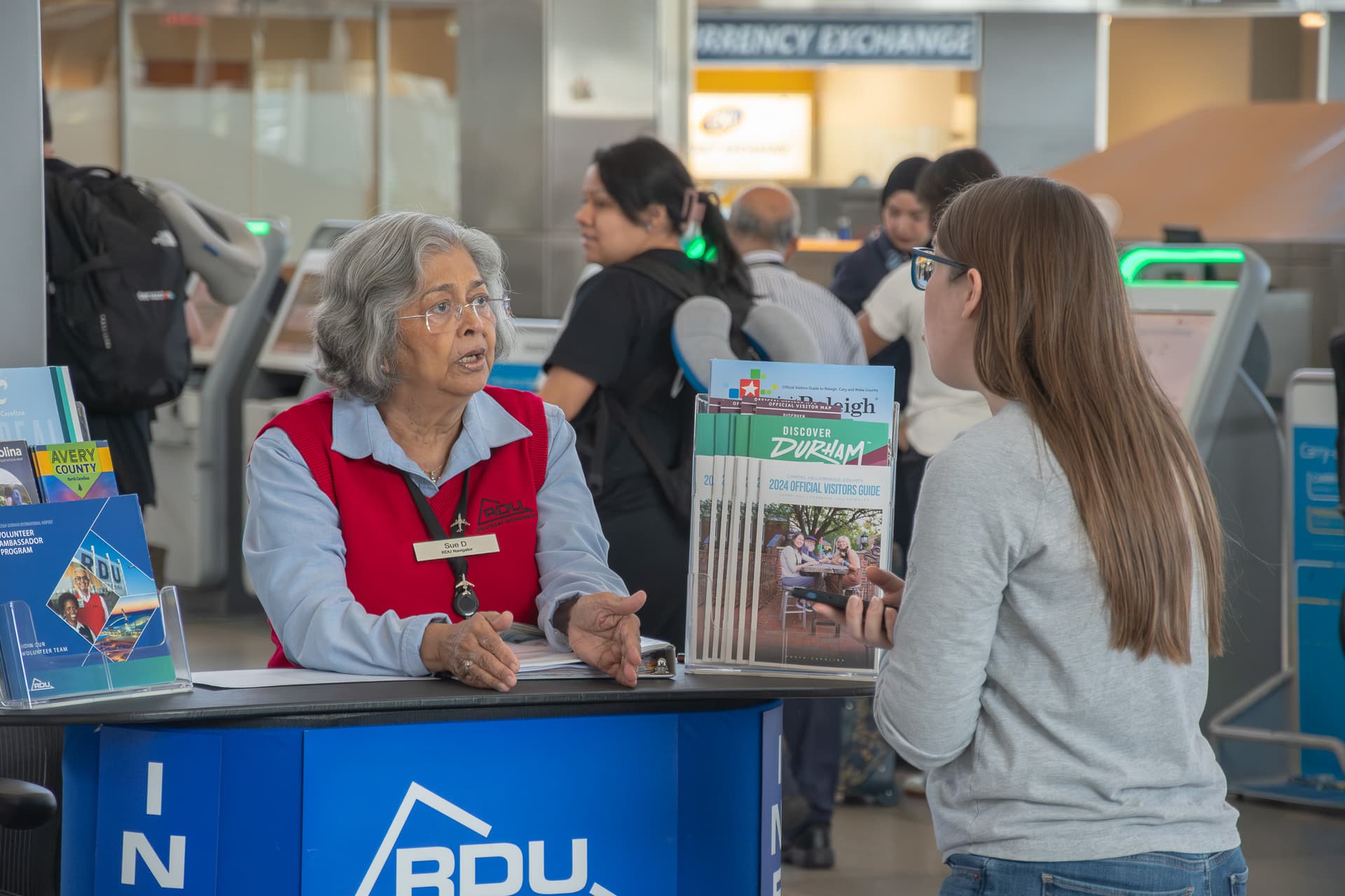 Airport volunteer in red vest assists traveler at information desk with Durham visitor guides displayed.