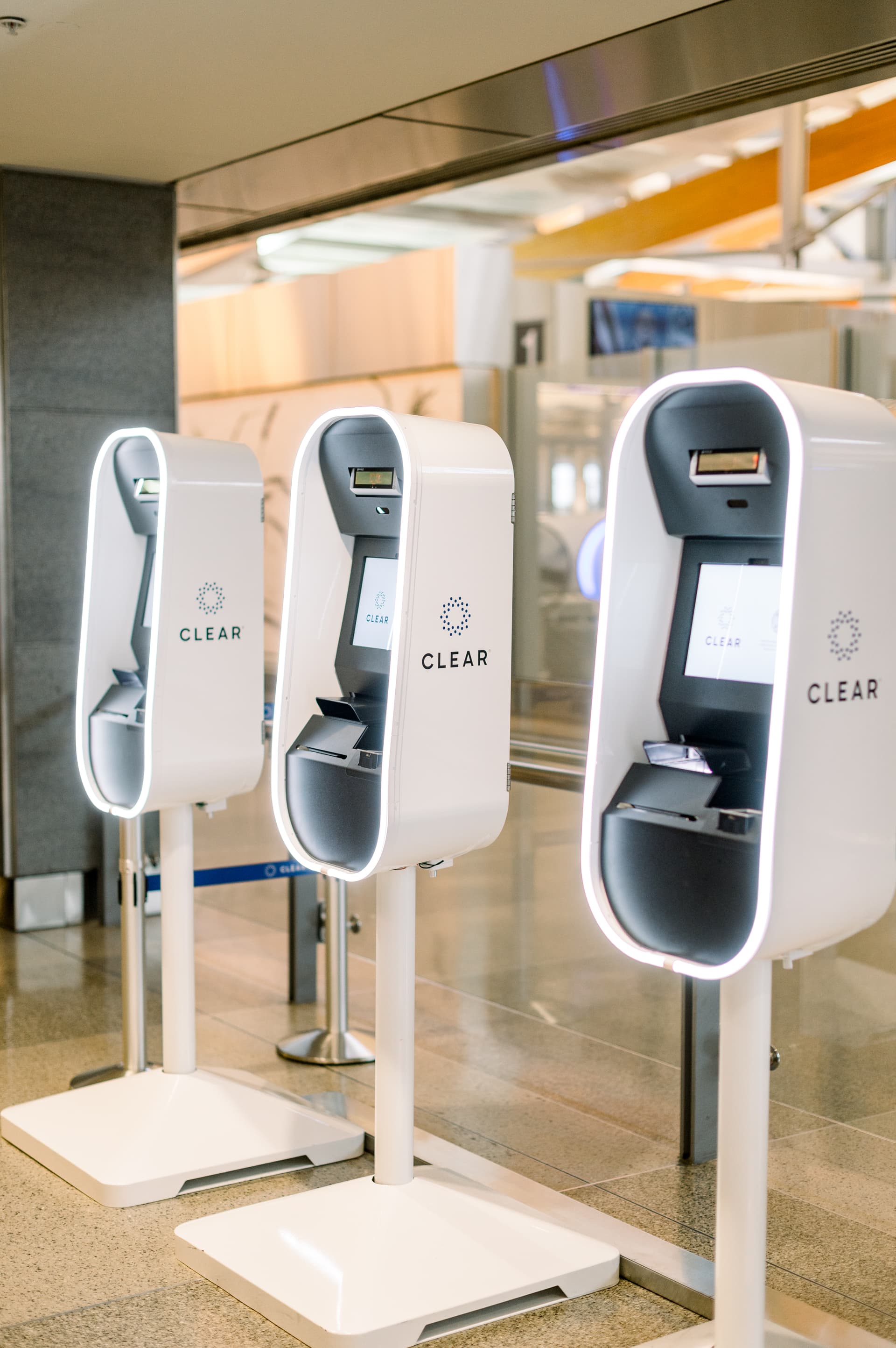 Three white CLEAR screening kiosks with gray scanners stand in a modern airport terminal with warm lighting.