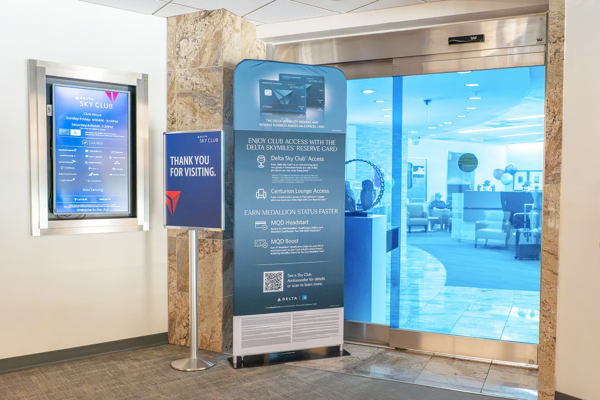 Delta Sky Club entrance with blue glass doors, digital screen showing hours and thank you sign with membership benefits.