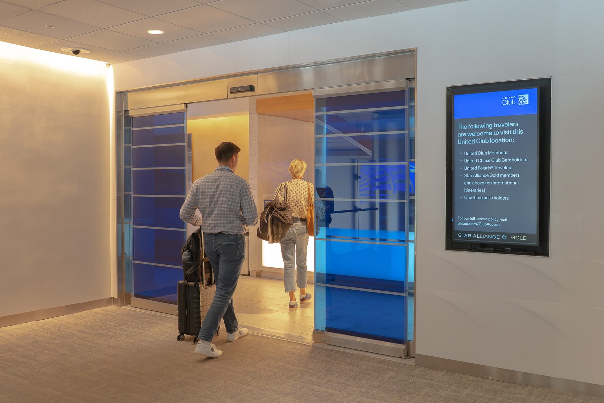 Two travelers with luggage entering United Club lounge through modern glass doors with blue accent panels and digital sign.