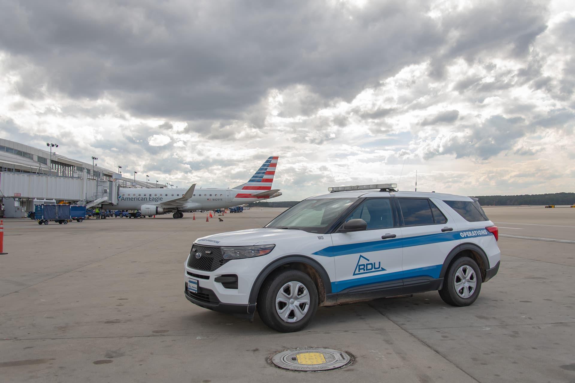 White airport operations SUV with blue stripes parked on tarmac with American Eagle plane and cloudy sky in background.