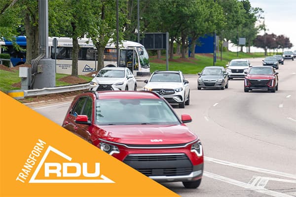 Red SUV leads traffic on tree-lined street with white vehicles following and orange Transform RDU logo in corner.