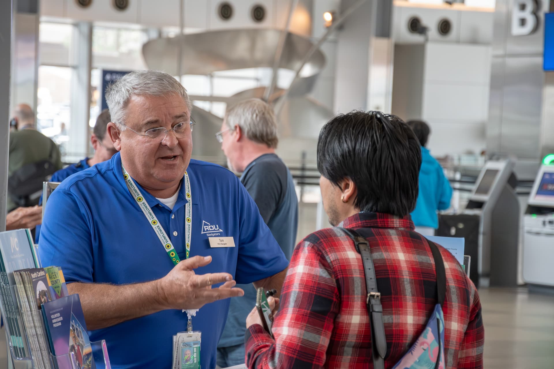 Staff member in blue polo shirt with lanyard speaking to visitor in red plaid shirt inside modern building lobby.