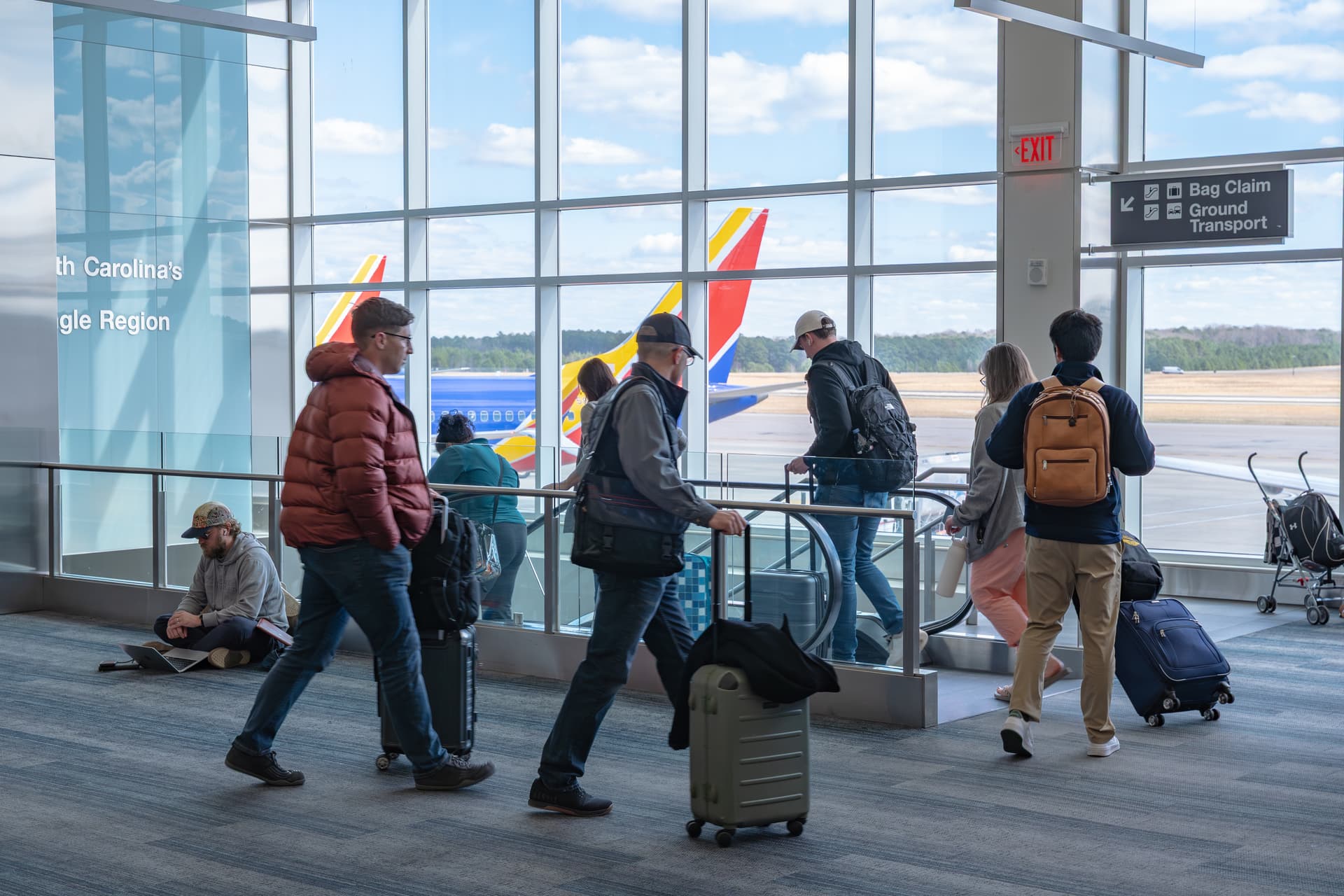 Travelers with luggage walking through modern airport terminal with large windows showing Southwest airplane on tarmac.