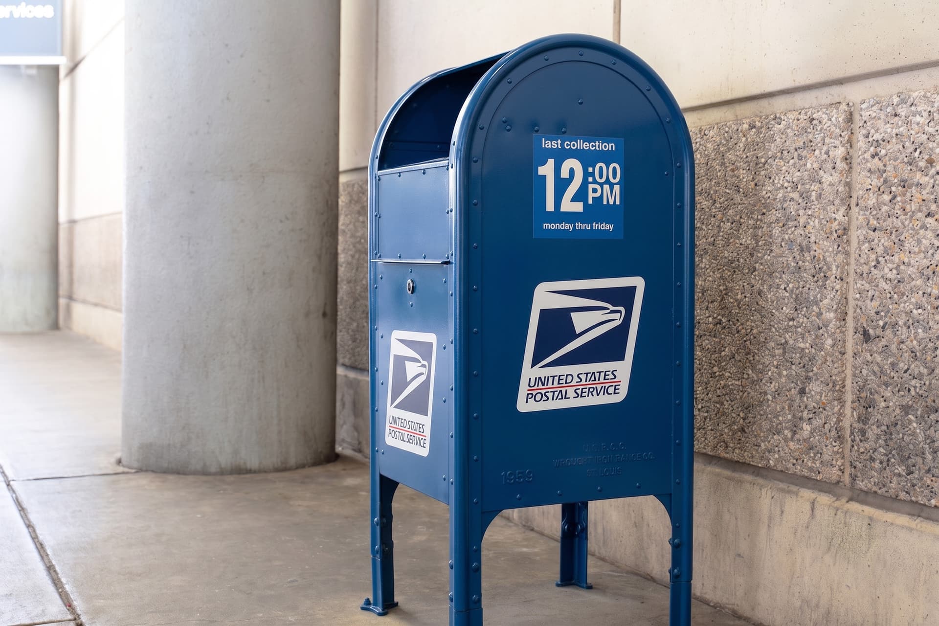 Blue USPS mailbox with last collection time of 12:00 PM Monday through Friday displayed on front panel.