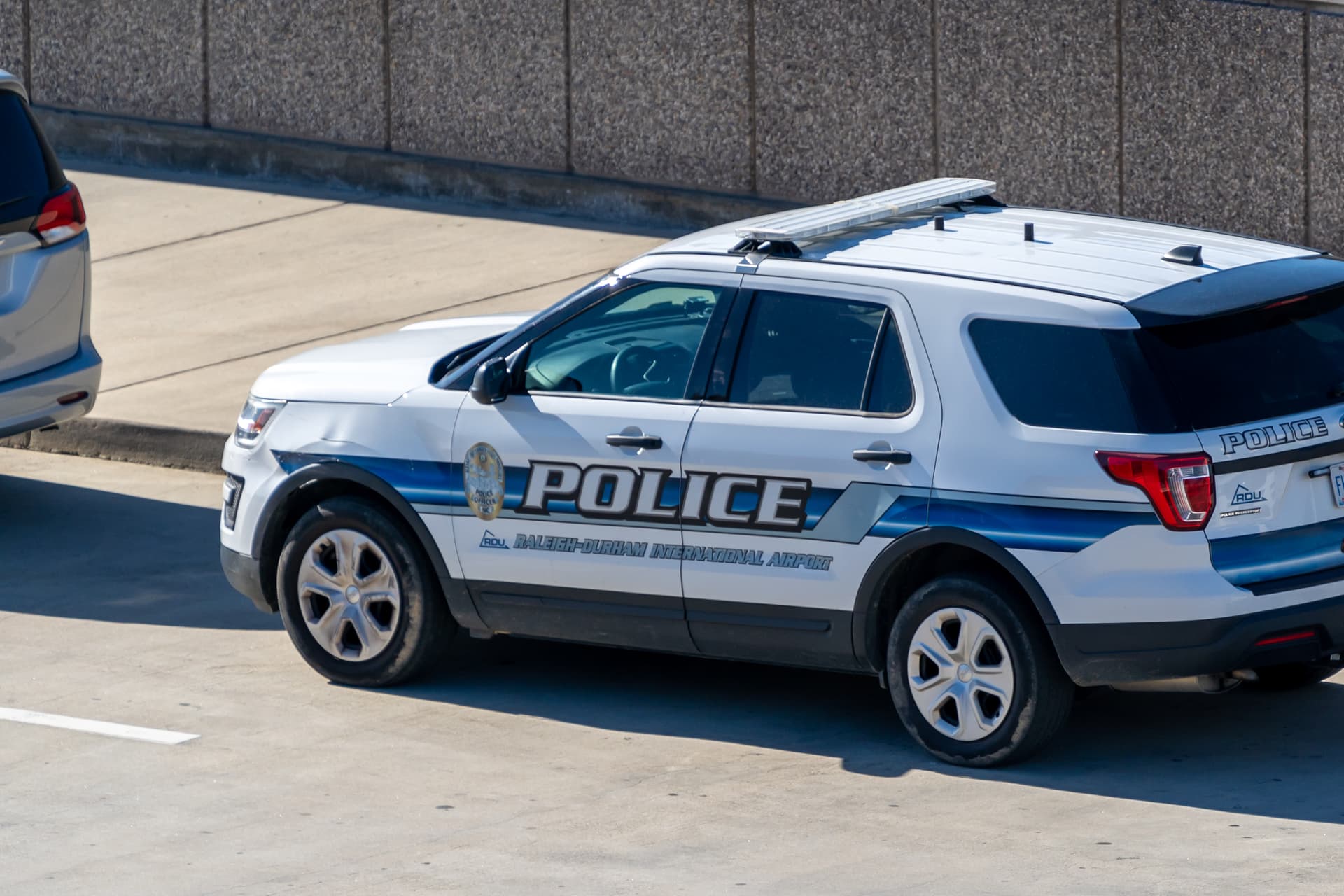 White Raleigh-Durham International Airport police SUV with roof rack parked on pavement near concrete wall.
