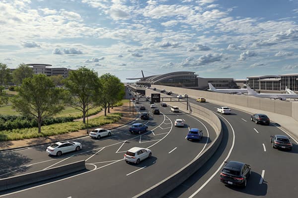 Aerial view of modern highway interchange with curved roads, vehicles, green landscaping, and commercial buildings under blue sky.