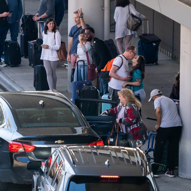 Travelers with luggage at airport pickup area, with couple embracing and cars waiting at curbside.