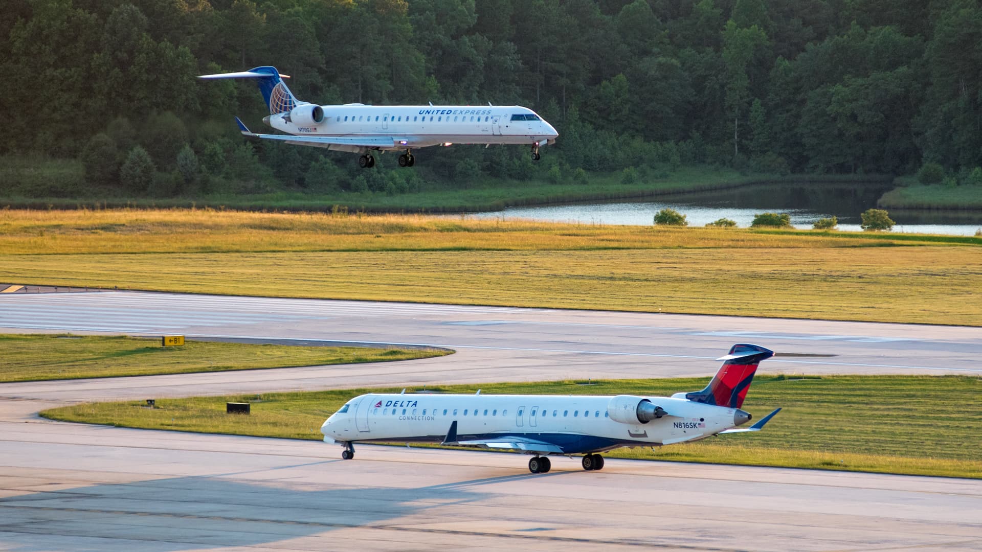 United Express jet landing above Delta aircraft on runway with trees and water in background.
