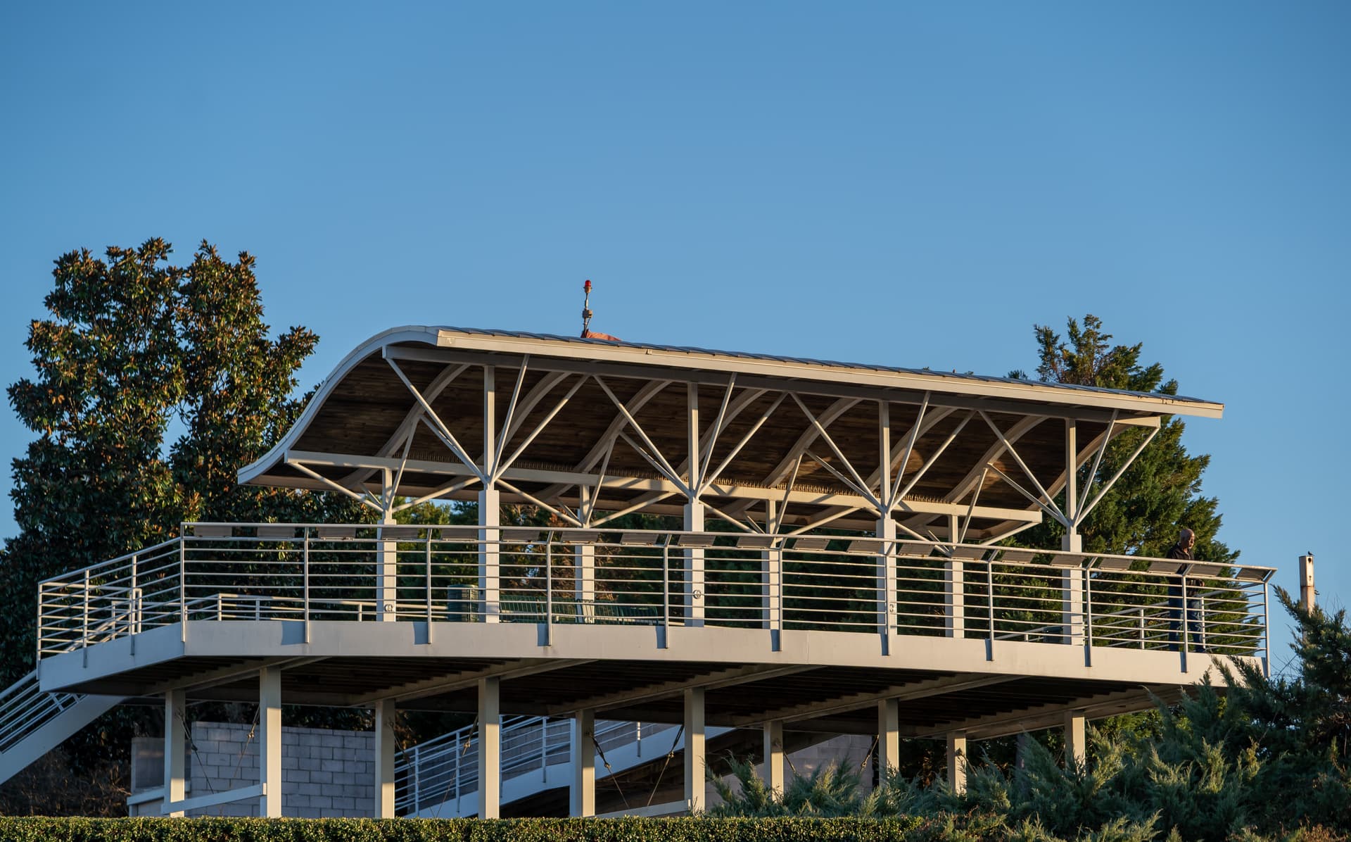 Modern pavilion with curved roof and diagonal support beams, elevated on pillars with metal railing, against blue sky.