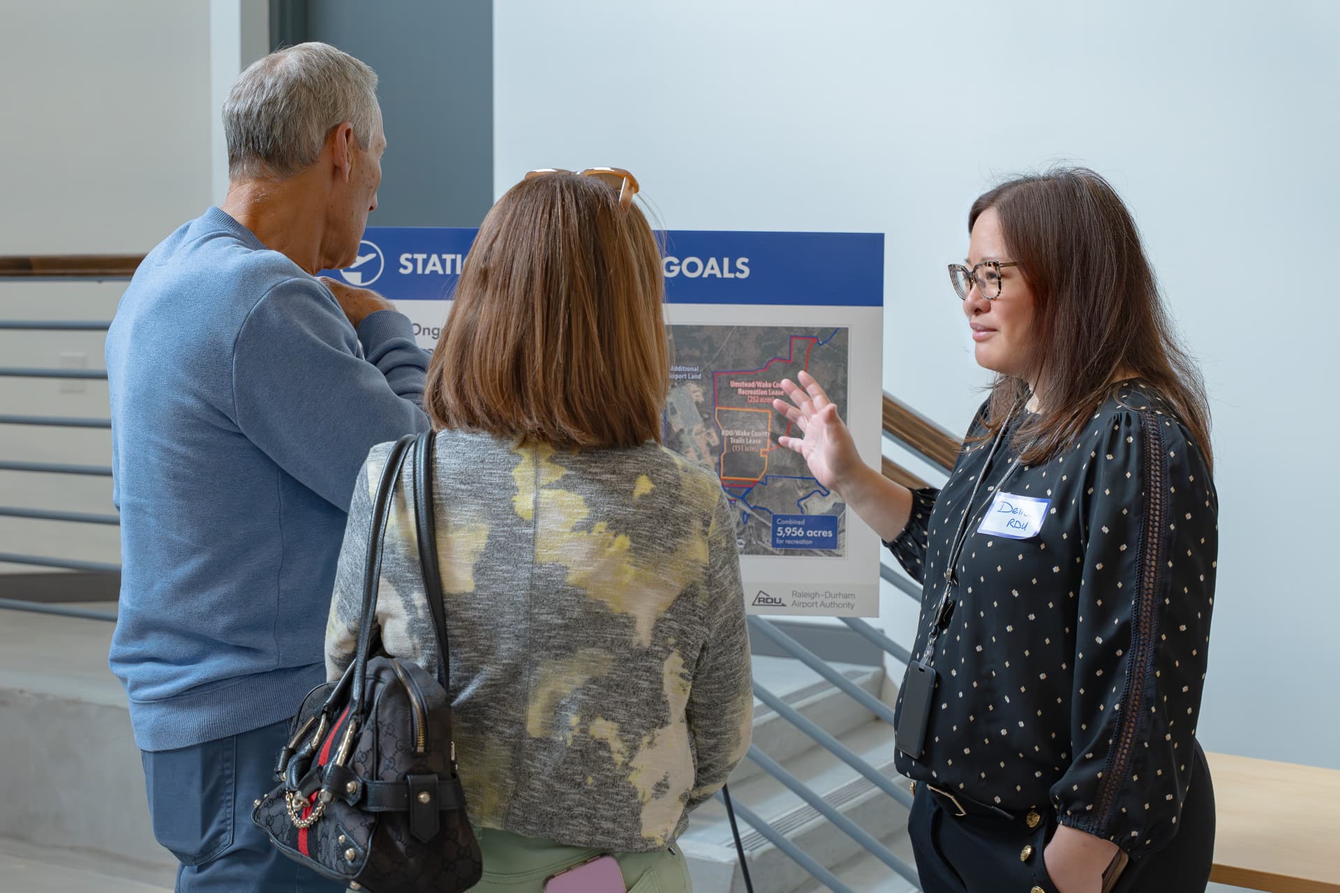 Woman in glasses presenting to two people in front of a blue banner displaying station goals with a map.