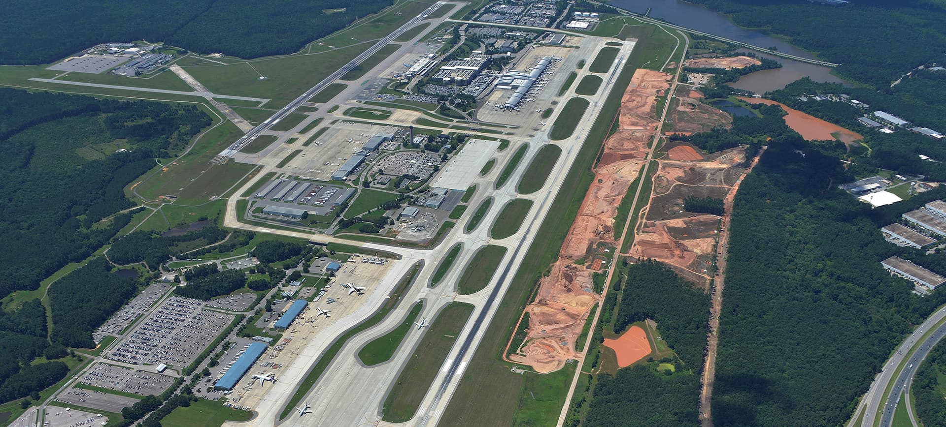 Aerial view of an airport with multiple runways, terminal buildings, parking lots, and adjacent red-orange construction areas.