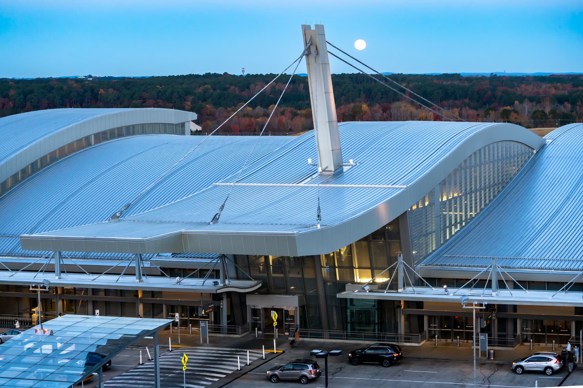 Modern airport terminal with curved metal roof and central tower against autumn forest and blue sky with moon visible.