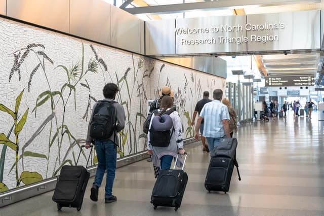 Travelers with luggage walking through airport terminal past botanical mural and Research Triangle Region sign.