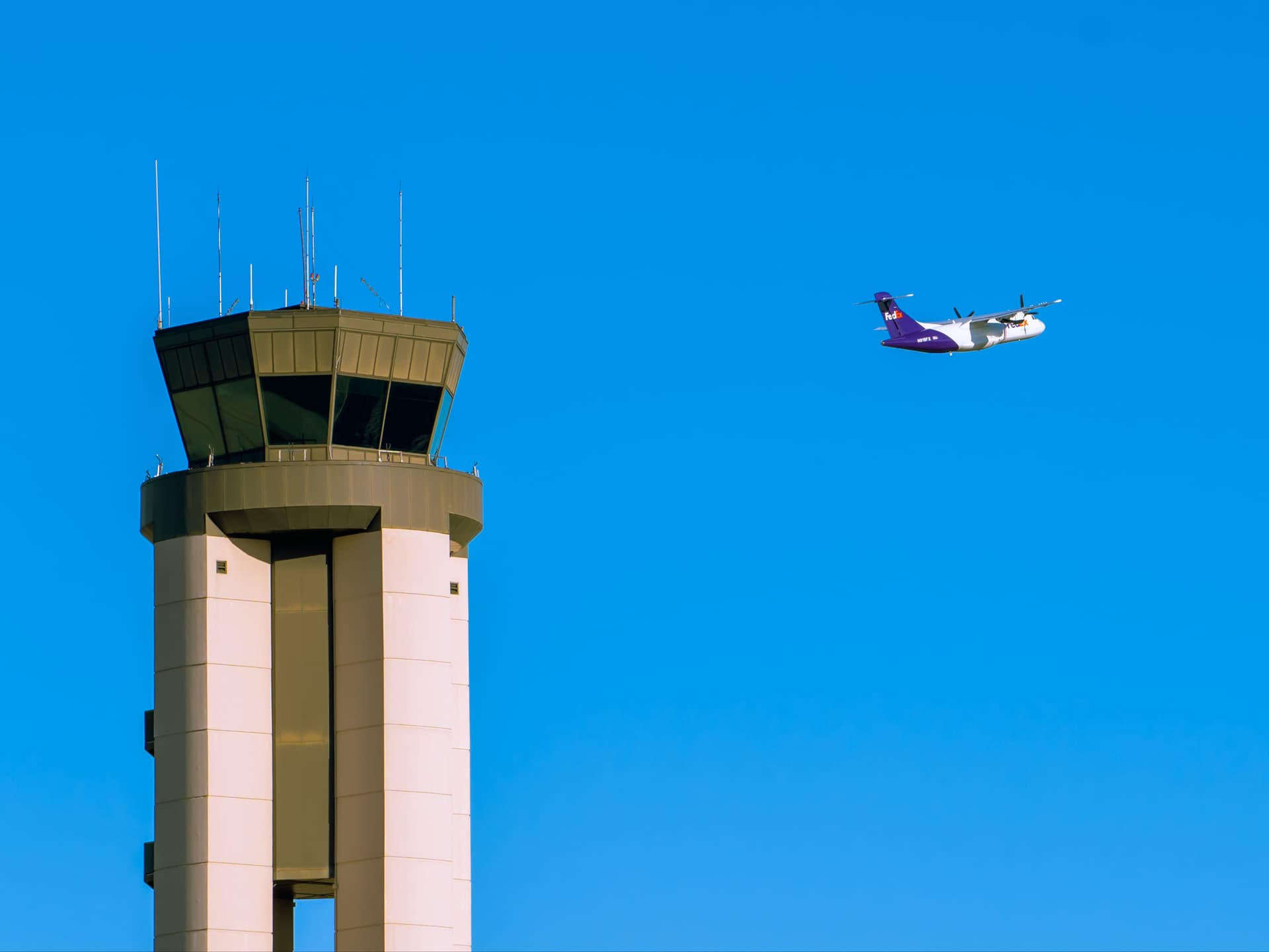 Purple and white plane flying past beige airport control tower against clear blue sky.