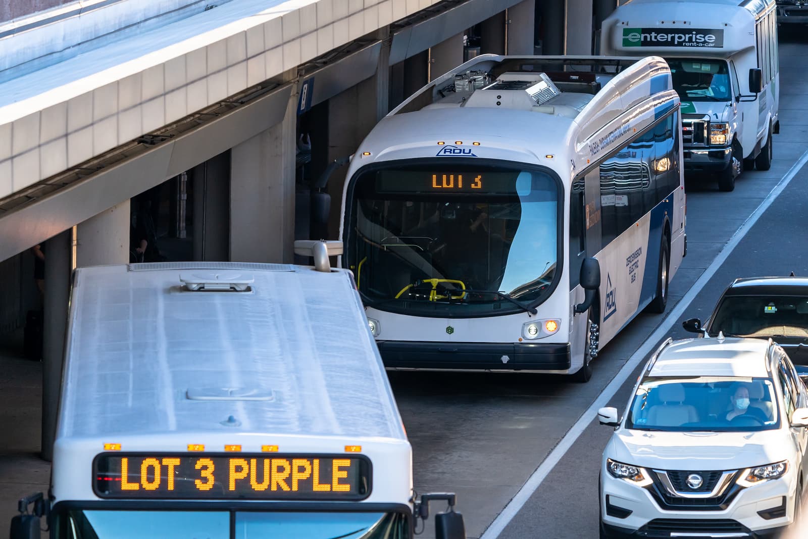 White airport shuttle bus displaying LUI 3 on digital sign, surrounded by vehicles on airport roadway near terminal.
